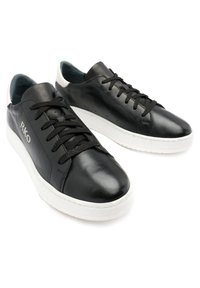 Baskets en cuir noir avec semelles en caoutchouc blanc, lacets noirs et texture lisse. Présentent des détails de logo et un design minimaliste.