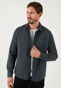 Camicia button-up grigio scuro in tessuto testurizzato, con taschino sul petto, design con colletto e maniche lunghe. Indossata su una maglietta bianca.