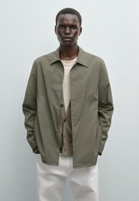 Olivengrøn letvægtsjakke med kravedesign, trykknapper, båret over en beige sweater og hvide bukser. Ensartet tekstur, afslappet pasform.