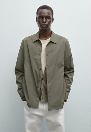 Olive green lightweight jacket with a collared design, snap buttons, worn over a beige sweater and white trousers. Plain texture, relaxed fit.