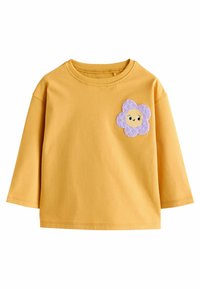 T-shirt jaune à manches courtes en coton. Présente un patch de fleur violette avec un visage souriant sur la zone gauche de la poitrine.