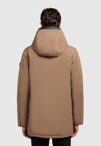 Brun vadderad jacka med huva, slät textur och krage prydd med logotyp. Har en avslappnad passform och sidofickor.