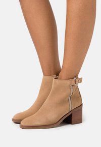 Botines de ante beige con puntera cuadrada y tacón cuadrado. Con cremallera lateral y una hebilla decorativa en el tobillo.