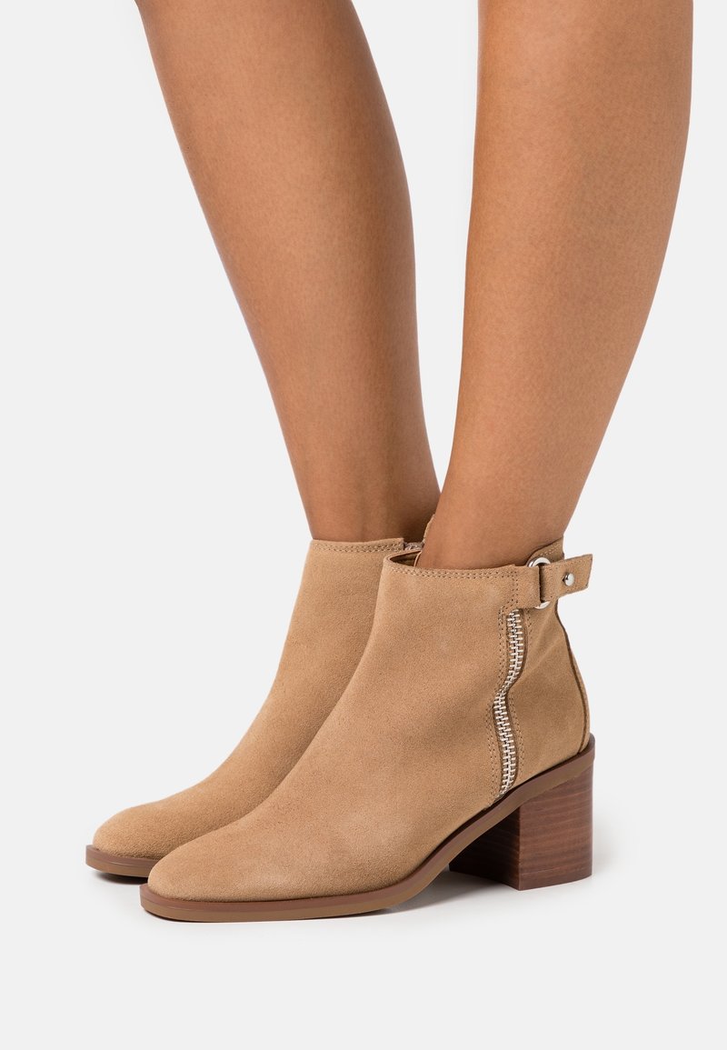 Botines de ante beige con puntera cuadrada y tacón cuadrado. Con cremallera lateral y una hebilla decorativa en el tobillo.