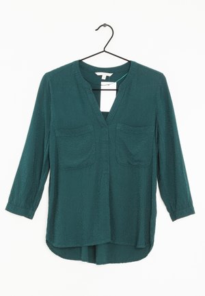 Blusa teal con maniche lunghe, scollatura a V, due tasche sul petto, tessuto testurizzato e orlo curvo. Appesa a una gruccia in metallo nero.