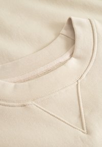 Beige sweatshirt with a ribbed crew neckline. Features a textured finish and a decorative stitched detail on the shoulder.