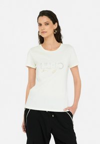 T-shirt blanc à manches courtes avec un design de logo pailleté sur le devant, fabriqué en tissu doux. Associé à un pantalon noir.