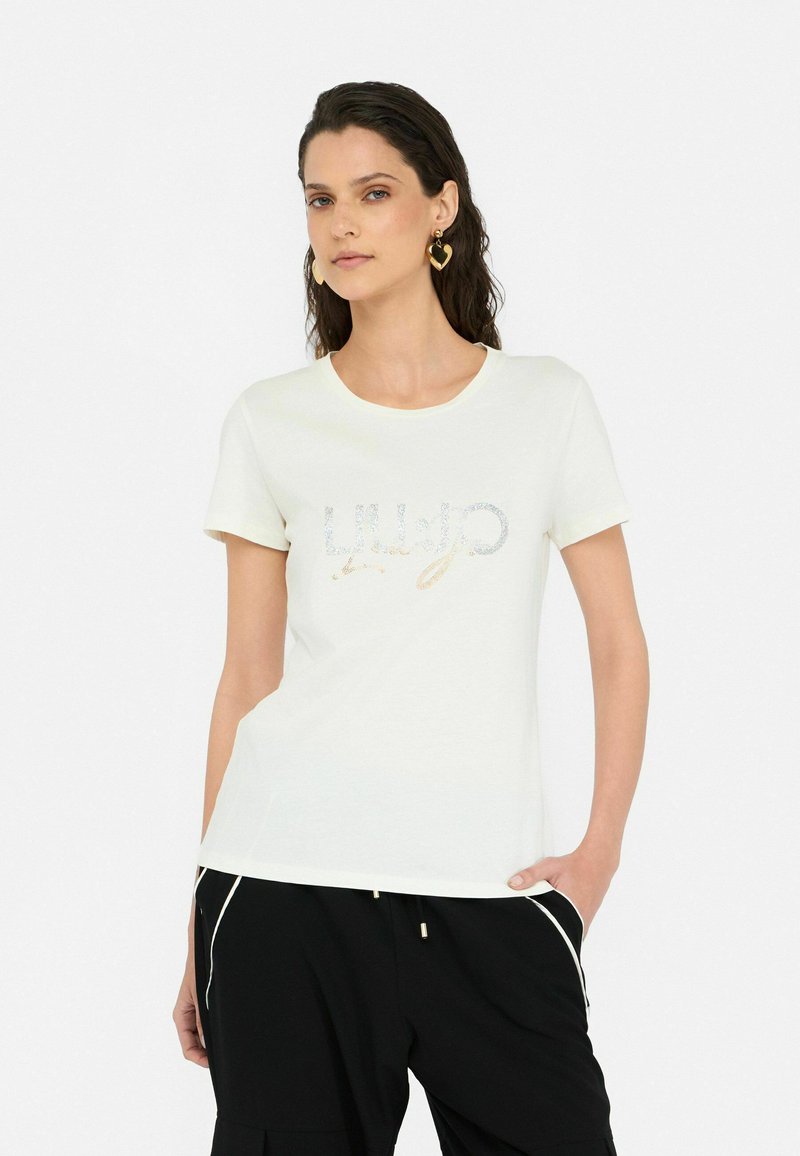 T-shirt blanc à manches courtes avec un design de logo pailleté sur le devant, fabriqué en tissu doux. Associé à un pantalon noir.