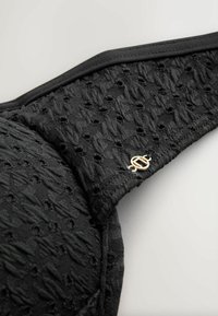 Bretelle de soutien-gorge texturée noire avec un petit logo circulaire doré sur un fond gris clair.