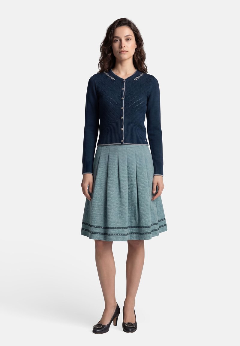 Femme portant un cardigan bleu marine boutonné avec broderie et une jupe plissée bleu clair longueur genou, debout sur un fond blanc.