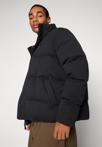 Jachetă puffer neagră cu textură matlasată și guler înalt. Dispune de un fermoar frontal, buzunare laterale și un branding subtil pe piept.