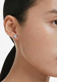 Detailný záber na tvár osoby, ktorá má v ľavom uchu náušnicu so modrým drahokamom a elegantné tmavé vlasy stiahnuté dozadu.