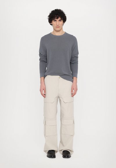 Graues Strickoberteil mit langen Ärmeln, kombiniert mit beigen Cargo-Hosen mit mehreren Taschen. Das Model steht vor einem schlichten weißen Hintergrund.