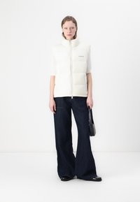 Hvit puffervest med høy krage, glidelås og logodetalj, kombinert med vide, mørke denimjeans. Enkel svart håndveske som tilbehør.
