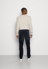 Beige Langarmshirt, dunkelblaue Jeans und weiße Sneaker. Das Shirt hat einen Rundhalsausschnitt; die Jeans haben Taschen hinten und einen geraden Schnitt.