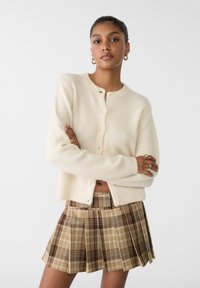 Cremefarvet uldblandings cardigan med rund halslinje og fem knapper, parret med en brun ternet plisseret nederdel.