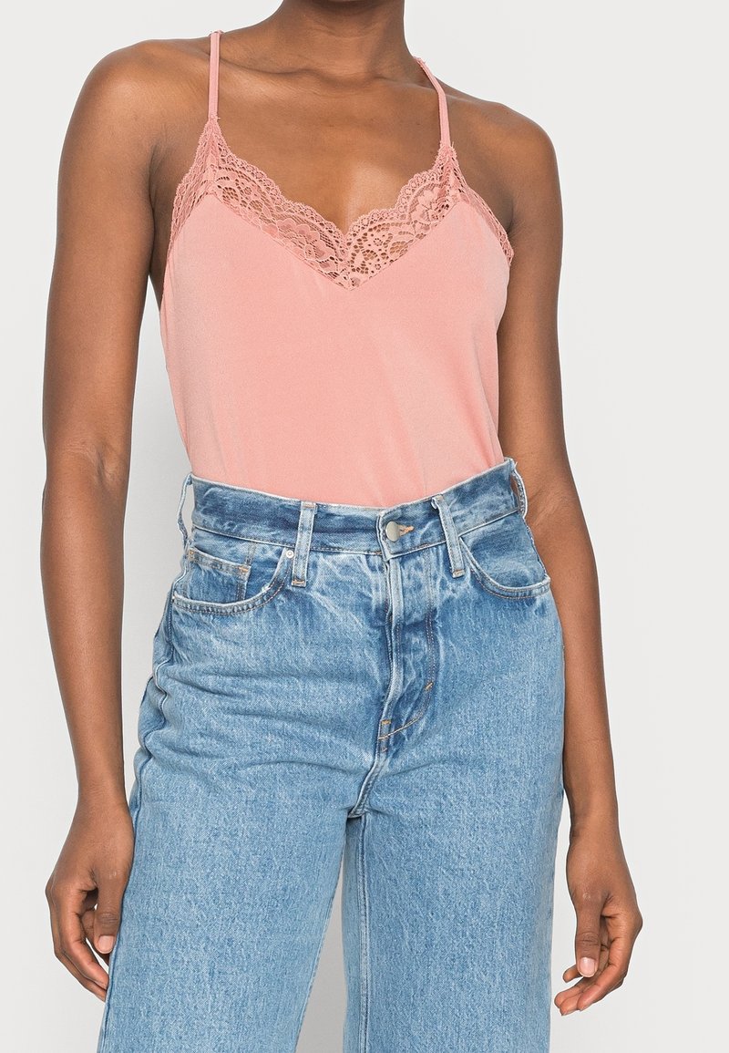 Femme portant un caraco rose bordé de dentelle rentré dans un jean denim bleu clair taille haute avec passants de ceinture et poches avant.