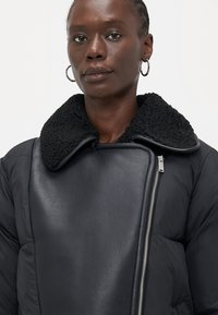 Persona che indossa una giacca nera con colletto in shearling testurizzato e zip asimmetrica, abbinata a orecchini a cerchio argentati.