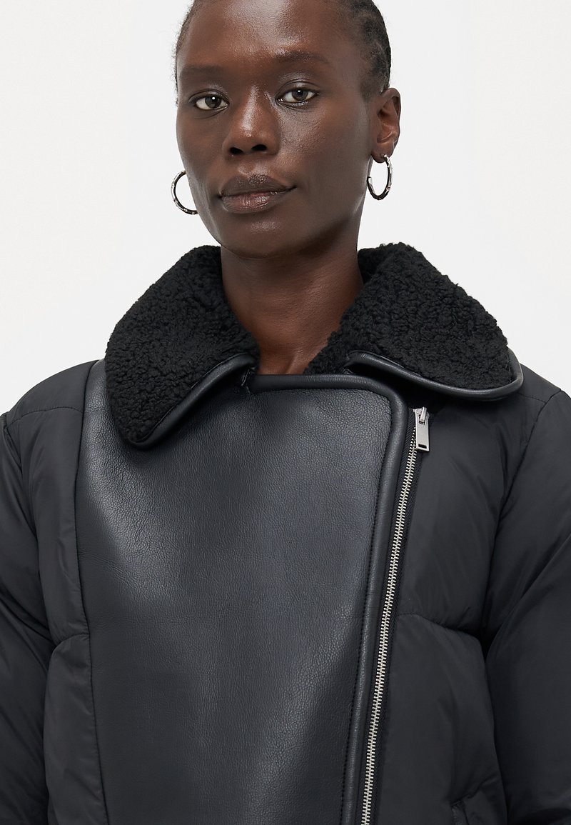 Persona che indossa una giacca nera con colletto in shearling testurizzato e zip asimmetrica, abbinata a orecchini a cerchio argentati.