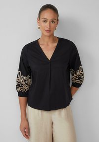 Zwarte blouse met een V-hals en driekwartmouwen, met gouden geborduurde bladmotieven op de mouwen. De stof is soepel en lichtgewicht.