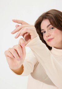 Langärmliges, geripptes Turtleneck-Oberteil in hellbeige; das Model posiert mit sanft berührenden Händen an dem Stoff und zeigt die Textur und Passform.