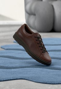 Baskets en cuir marron avec surface texturée, bout rond et lacets noirs. Semelle légère et détail de couture subtile. Flottant au-dessus d'un tapis bleu.