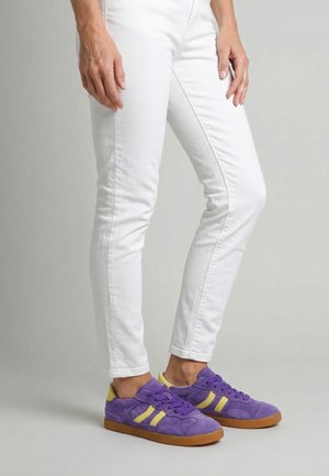 Osoba nosząca białe obcisłe spodnie i fioletowe trampki z żółtymi paskami i gumową podeszwą, stojąca na jasnoszarym tle.