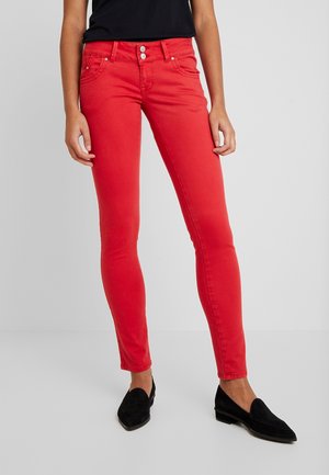 Strakke rode broek gedragen met zwarte loafers en een ingestopt zwart topje, gedragen door een staande persoon tegen een effen grijze achtergrond.