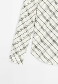 Chemise blanche à manches longues avec un motif à carreaux écossais noir et gris, montrant la manche et la partie basse du torse.