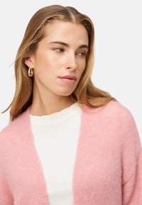 Lichtroze, pluizig vest met open voorkant, gedragen over een wit shirt. Heeft een zachte textuur en een ontspannen pasvorm. Goudkleurige oorbellen in de vorm van hoepels.