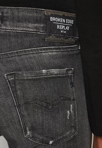 Vaqueros de mezclilla negra con una textura desgastada, que presentan un bolsillo trasero con detalles de costura y una etiqueta que dice "Broken Edge Replay."