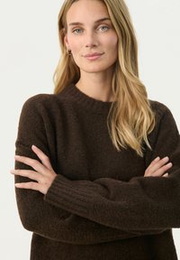 Hnědý pletený svetr s ribovaným výstřihem a manžetami. Měkká, texturovaná tkanina. Model s překříženýma rukama, ukazující uvolněný střih a detaily designu.