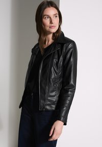 Veste en cuir noir de motard avec poches zippées, col cranté et détails cannelés sur les manches, portée sur un haut noir.