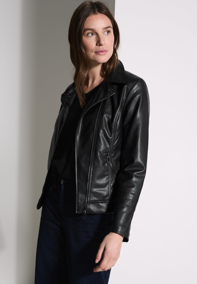 Veste en cuir noir de motard avec poches zippées, col cranté et détails cannelés sur les manches, portée sur un haut noir.