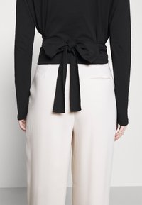 Schwarzes Langarmoberteil mit geknotetem Rückendetail, kombiniert mit cremefarbener, weit geschnittener Hose, glattes Stoffgefühl und minimalistisches Design.
