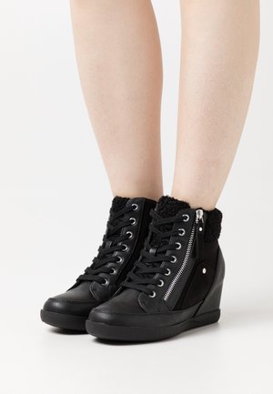 Bottines compensées noires avec lacets, fermeture éclair latérale et col texturé, portées sur des jambes nues sur fond blanc.