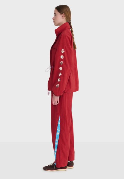 Femme en veste rouge et pantalon avec des motifs pixellisés blancs sur la manche et une bande bleue avec des motifs blancs sur la jambe du pantalon, debout de profil.