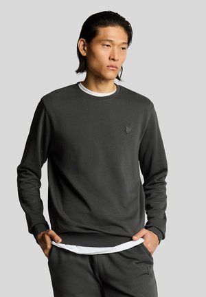 Mand med sort hår iført en mørkegrå sweatshirt og matchende bukser med hænderne i lommerne, stående mod en ensartet lys grå baggrund.