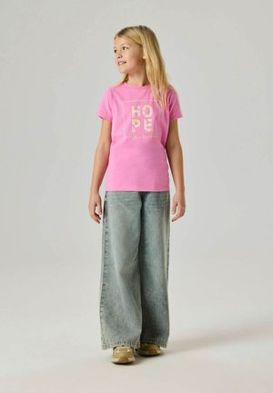 Młoda dziewczyna o długich blond włosach, ubrana w różowy T-shirt z napisem "HOPE", szerokie szare jeansy i beżowe trampki, patrząca w bok i uśmiechająca się.