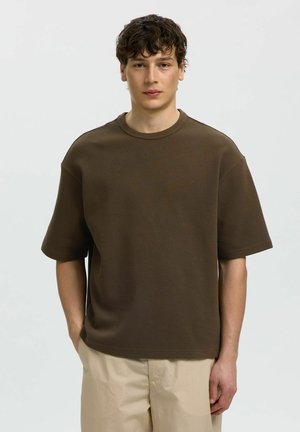 Junger Mann mit kurzen lockigen Haaren, der ein locker sitzendes braunes texturiertes Kurzarmhemd und beige Hose trägt, steht vor schlichtem Hintergrund.