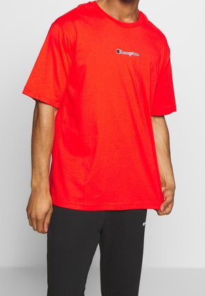 Person iført en skrigrød Champion T-shirt og sorte bukser, der holder i kanten af T-shirten med højre hånd.