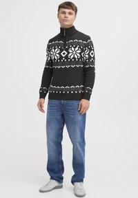 Svart genser med hvitt snøkrystallmønster, høy krage med glidelås, finstrikket tekstur. Brukt med blå jeans og grå sneakers.