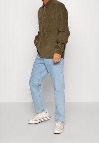 Olivově zelená kordurová košile na zip s náprsní kapsou, kombinovaná s lehce modrými džíny se rovnými nohavicemi a béžovými vysokými teniskami.