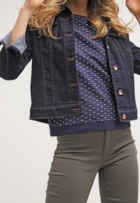 Kobieta w ciemnej dżinsowej kurtce, nosząca granatowy sweter w kropki oraz oliwkowe spodnie, pozująca z uniesionymi rękami.