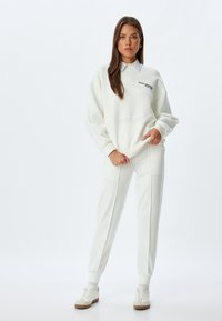 Felpa bianca con collo a costine, indossata sopra una camicia a righe con colletto, abbinata a pantaloni tapered coordinati. Entrambi i capi presentano una texture liscia.