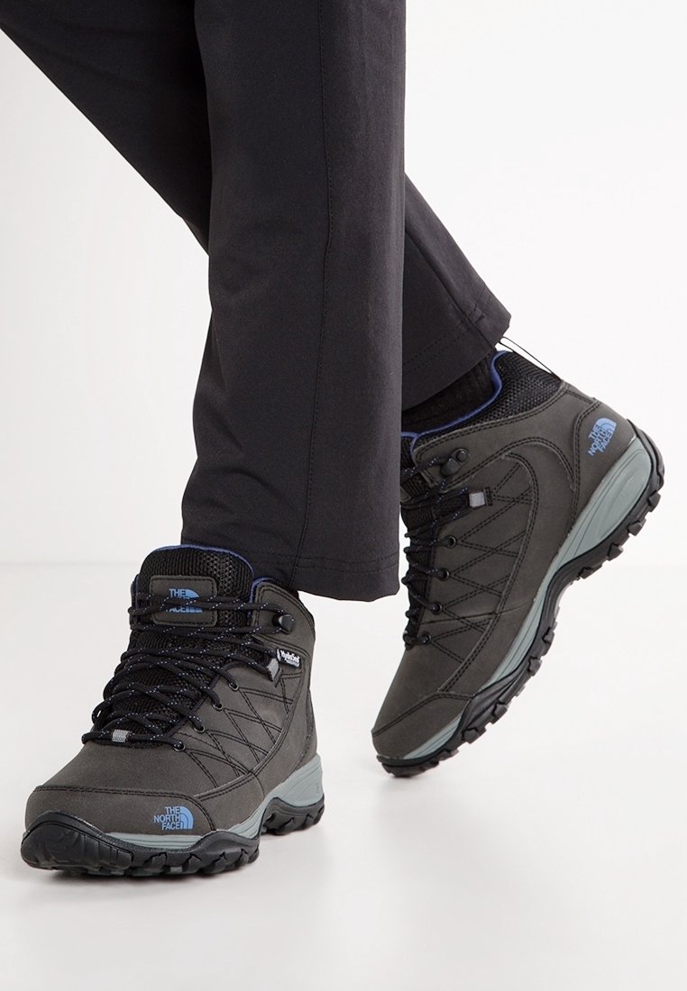 Personne portant des chaussures de randonnée noires avec des accents bleus et un pantalon noir, un pied légèrement levé sur un fond blanc uni.