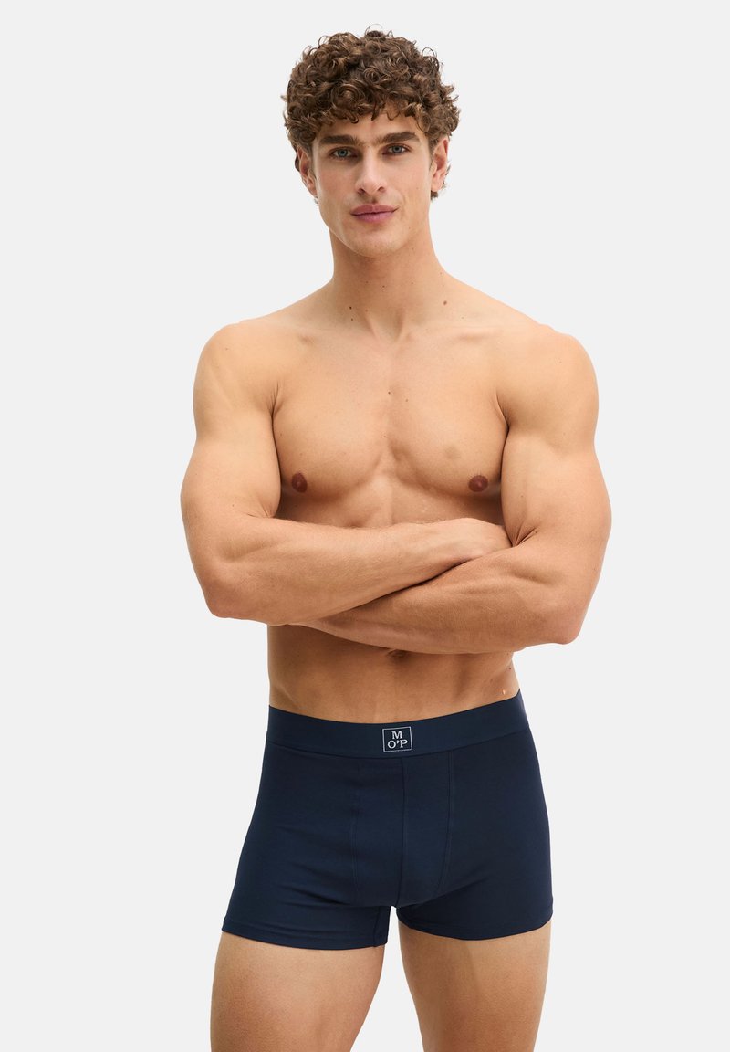 Muskulöser Mann mit lockigem Haar steht mit verschränkten Armen da, trägt dunkelblaue Boxershorts vor einem schlichten hellen Hintergrund.