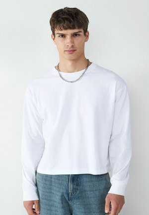 Weißes Langarmshirt mit Rundhalsausschnitt, im lässigen Schnitt. Kombiniert mit hellblauen Jeans. Akzentuiert mit einer silbernen Kette.