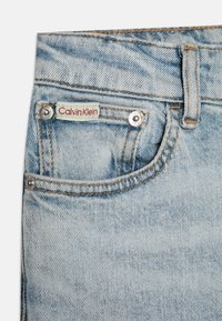 Lys blå denimjeans med en falmet finish, som har en rød "Calvin Klein"-etikett og sølvfarget detaljering, inkludert en knapp og nitter.