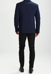 Marineblå kostymjacka med strukturerade axlar, parat med slim-fit svarta byxor. Slät tygtextur och knappar på ärmarna. Svarta skor.